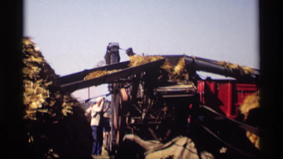 Jordan, Minnesota 1969 footage frame with workers near a grain processing machine and hay or grain piled on moving equipment.