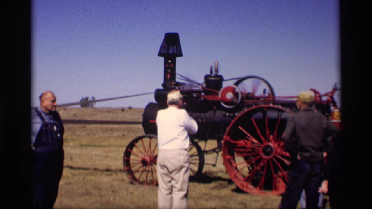 In Jordan, Minnesota, 1969, adults observe a red-and-black steam-powered tractor in an open field under a clear sky.