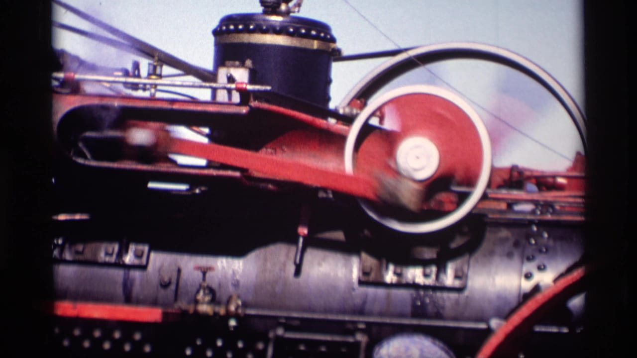 Close view of a steam tractor engine with a red connecting rod and spinning flywheel.