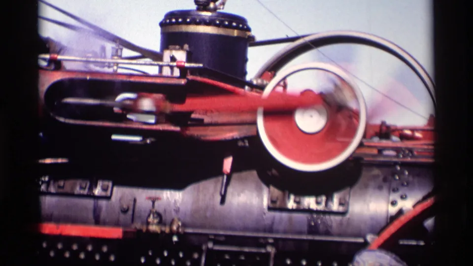 Tight 8mm view of the red-and-black steam tractor mechanism, with the flywheel blurred by motion against the boiler.