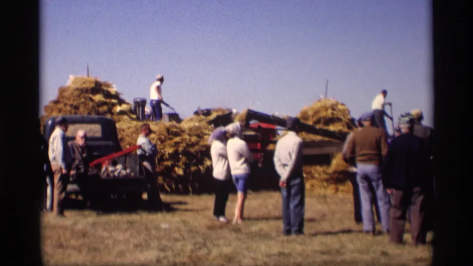 Wide home-movie view of a 1969 Jordan, Minnesota haying scene with spectators, workers, a truck, and machinery under a clear sky.