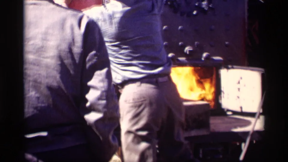 A handheld close-up shows two adults near the bright opening of an incinerator or firebox during the 1969 Jordan, Minnesota farm scene.