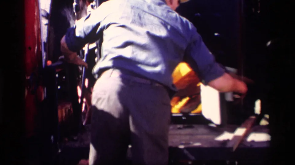 A man in work clothes bends toward a bright incinerator or firebox opening, suggesting hands-on tending during rural farm cleanup activity.