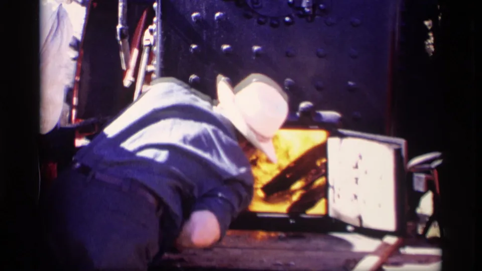 A worker in a light shirt and white hat tends the glowing steam locomotive firebox in Jordan, Minnesota, 1969.