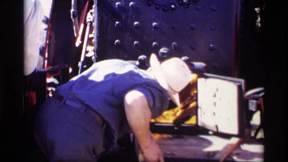 The 1969 home-movie frame shows a worker crouched at a steam train furnace with firelight reflecting on nearby metal.