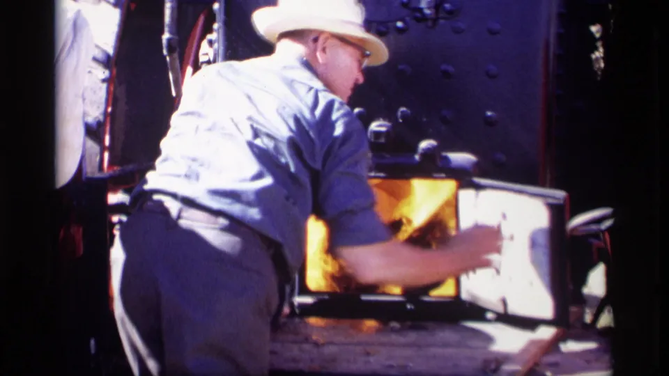A worker turns at the open steam locomotive firebox, his face partly visible against dark riveted metal and bright flames.