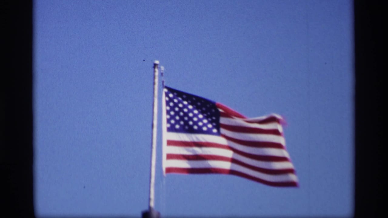 U.S. flag waving in bright sunlight against a blue sky in 1969 Jordan, Minnesota home-movie footage.