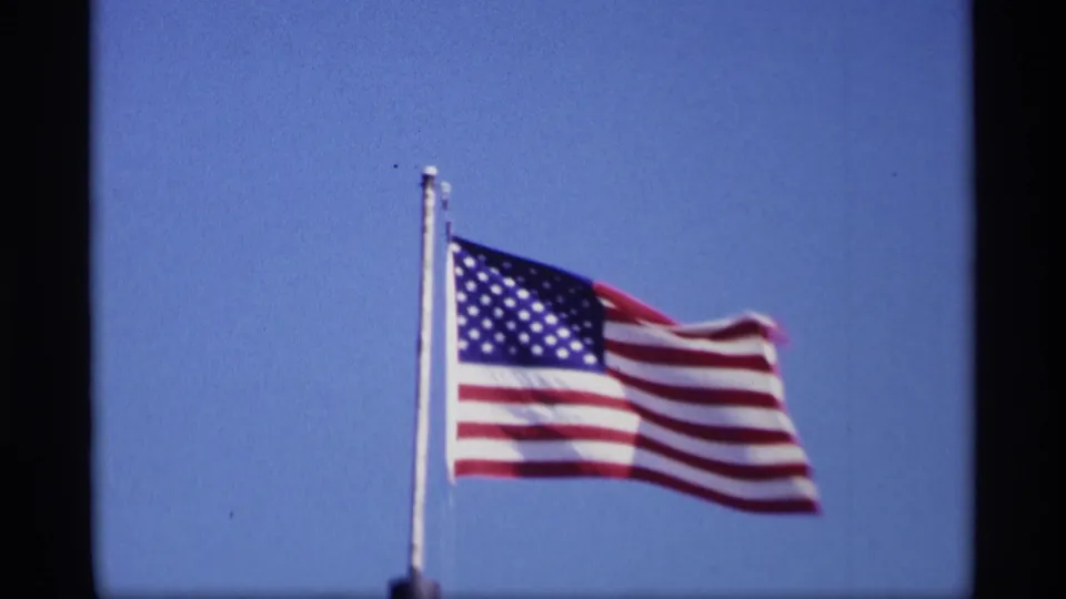 U.S. flag waving in bright sunlight against a blue sky in 1969 Jordan, Minnesota home-movie footage.