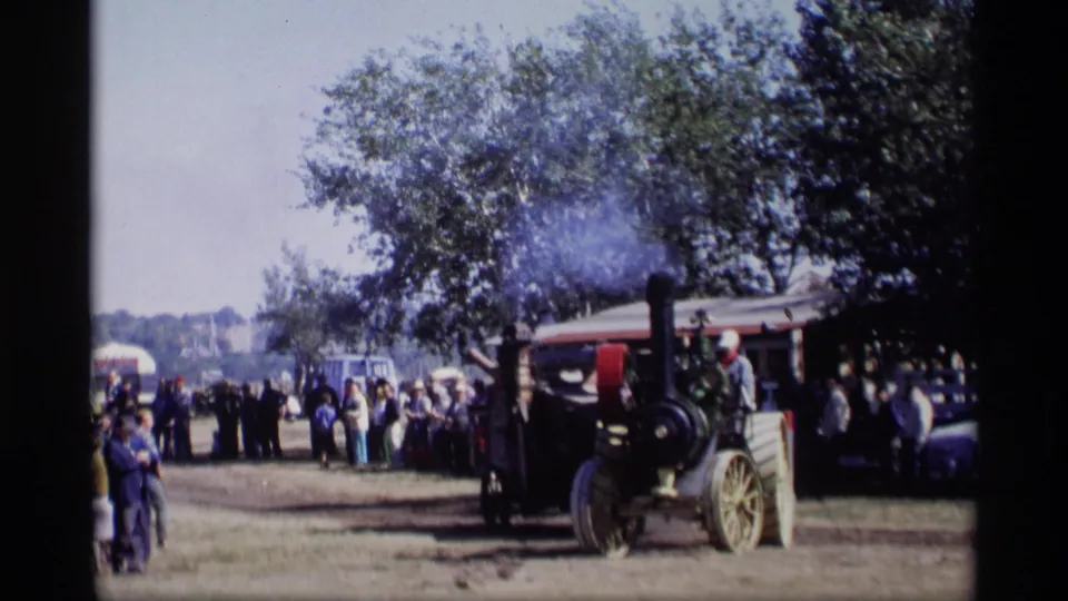An outdoor gathering in Jordan, Minnesota shows people near trees, parked vehicles, and a steam tractor emitting smoke.