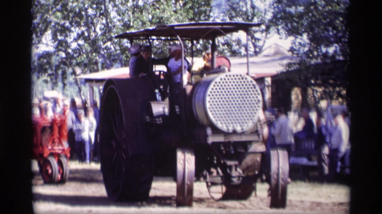 Steam tractor viewed from behind with passengers and a crowd in a grassy field.