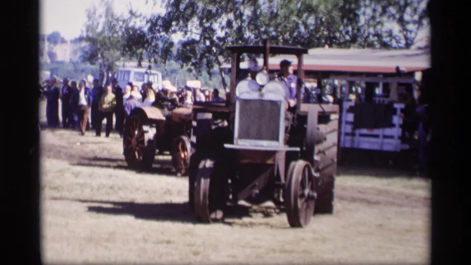 Front-facing tractor approaches across a grassy fairground in Jordan, Minnesota, with onlookers and another tractor nearby.