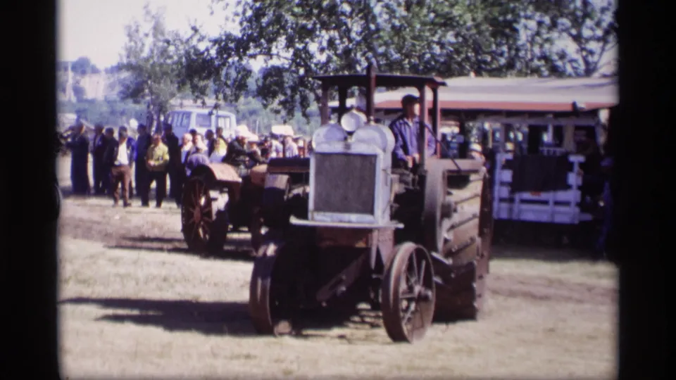 Home-movie frame from Jordan, Minnesota showing a tractor near the camera while a fairground crowd and another tractor remain behind it.