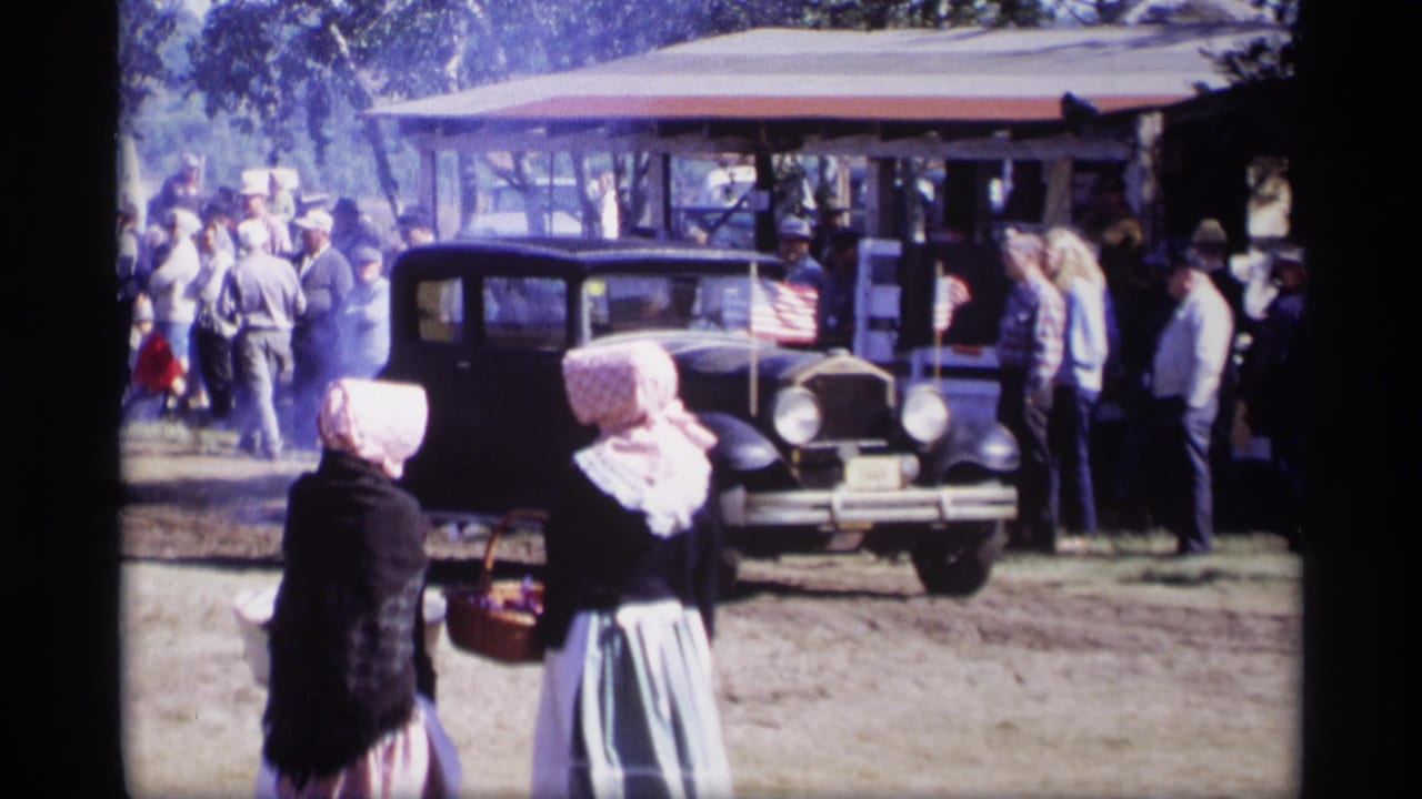 Crowd gathers around a black vintage car near a pavilion while two women stand in front.