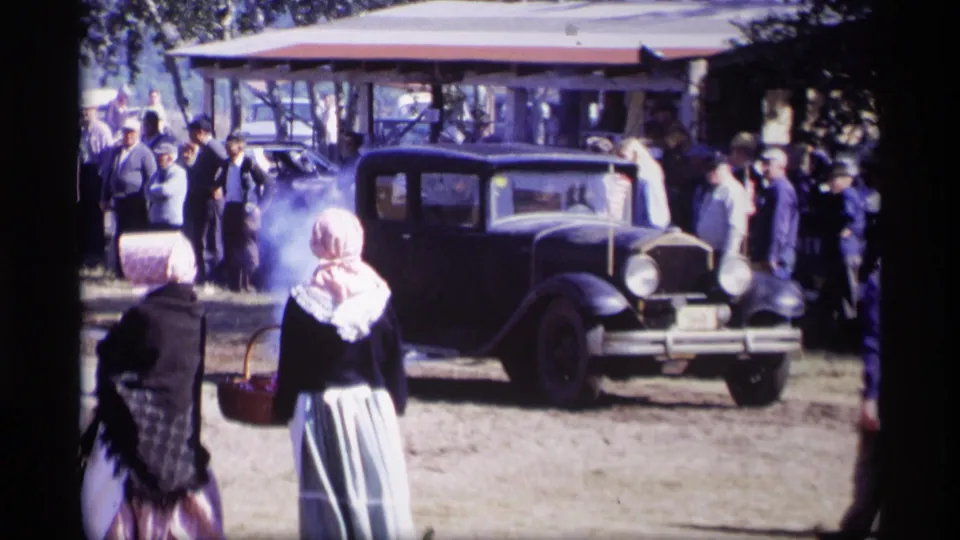 A black vintage car faces forward on dirt ground at the 1969 Jordan, Minnesota event, with bonneted women and a nearby crowd partly obscured by haze.