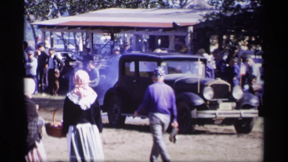 A man crosses the foreground in a 1969 Jordan, Minnesota home-movie shot as a black vintage car and pavilion crowd remain behind him.