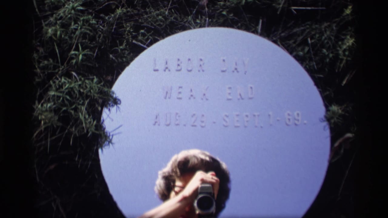 Person partly visible holding a camera below a white circular Labor Day weekend title card in grass.