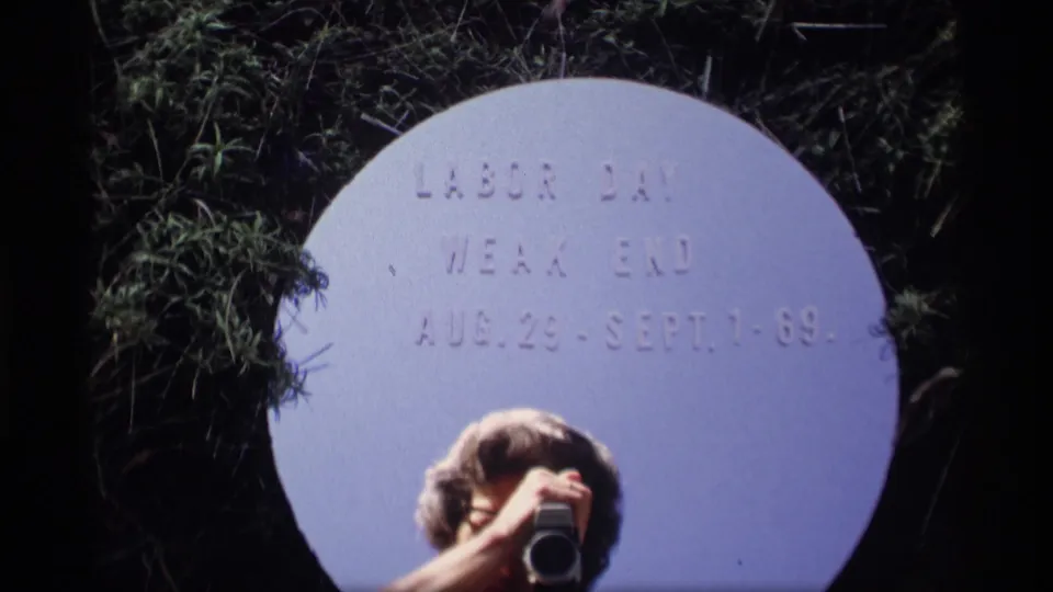 1969 home-movie title setup with Labor Day weekend dates on a white circular card and a camera held by a partly visible person.