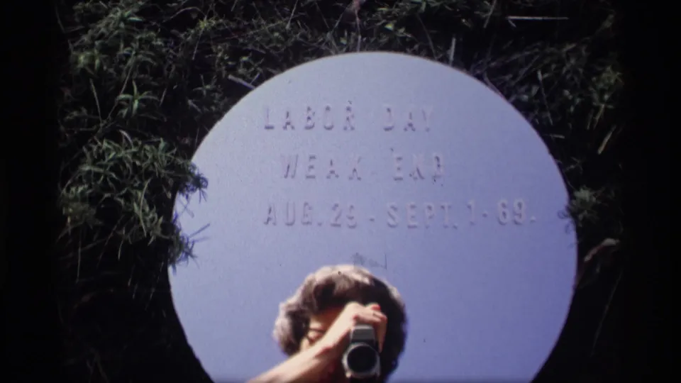 A white circular Labor Day weekend title card dated Aug. 29-Sept. 1-69 is framed with a partly visible person and camera below.