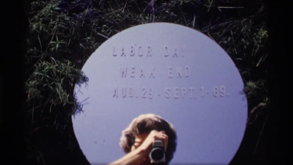 1969 home-movie frame with a circular Labor Day weekend sign and a person with glasses holding a camera in the foreground.