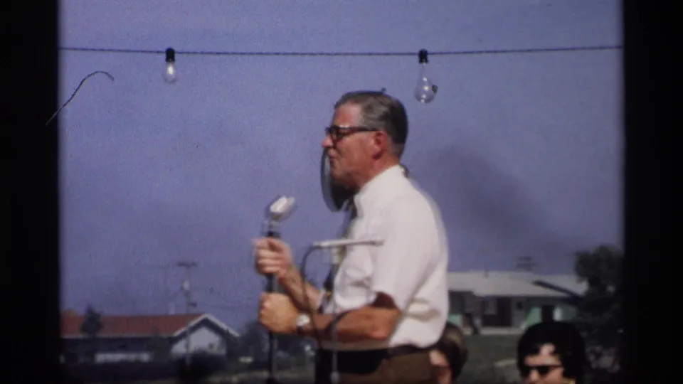 In Mabel, Minnesota, 1969, a man in a white shirt and tie appears to speak into a microphone at an outdoor gathering.