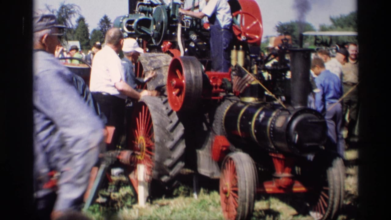 Crowd gathered around a steam tractor with red wheels.