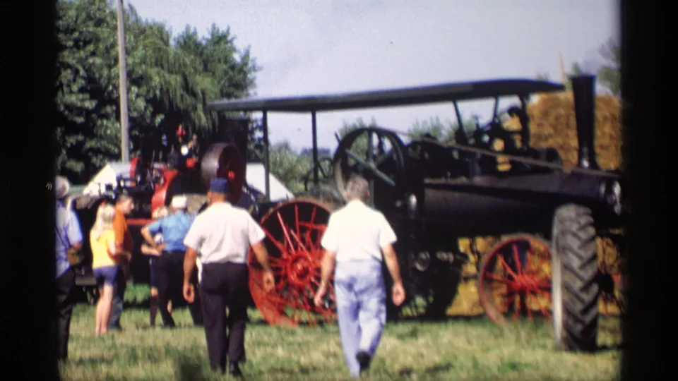 1969 Mabel, Minnesota 8mm frame of visitors walking beside a preserved steam tractor with red wheels at a rural gathering.
