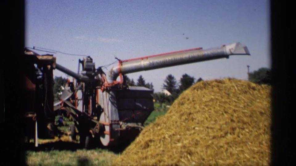 1969 Mabel, Minnesota 8mm frame of a thresher working near a straw pile, with trees and distant farm structures behind it.