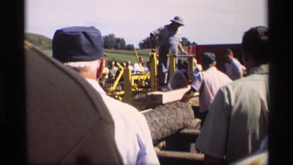 From within the crowd, this 1969 Mabel, Minnesota frame shows spectators watching a yellow wood-cutting machine and operator near logs.