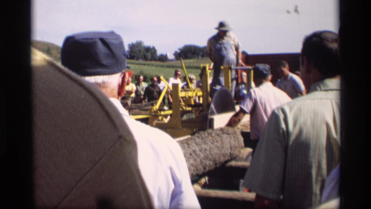 1969 Mabel, Minnesota 8mm frame with spectators facing a man near a yellow saw rig and a long piece of lumber in a rural field setting.