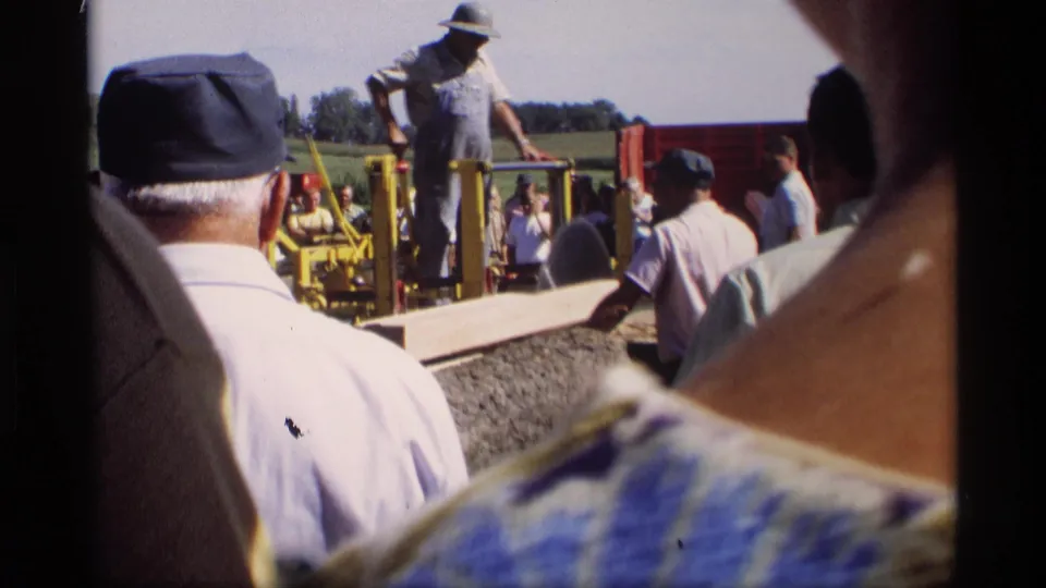 A crowd in casual work clothes watches a man at a yellow industrial saw during the 1969 rural woodworking scene in Mabel, Minnesota.