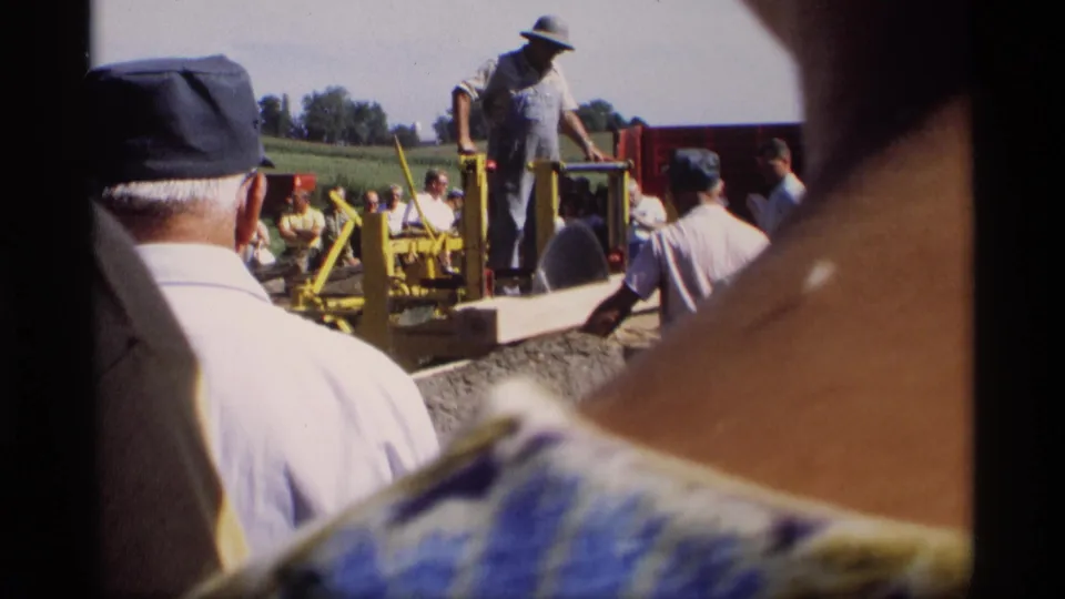 Handheld 8mm view partly blocked by nearby spectators as the saw operator stands over a long board at the Mabel, Minnesota demonstration.
