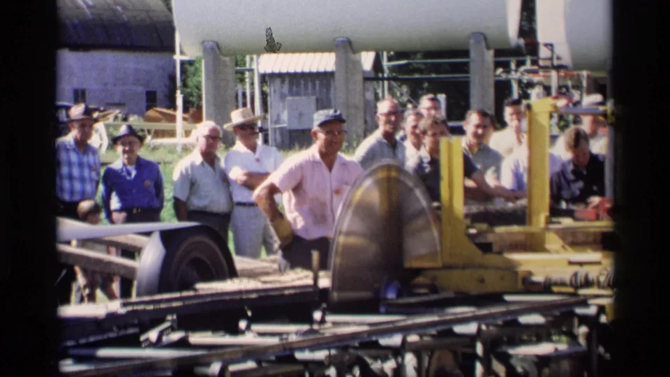 Large circular saw blade, yellow machinery, and assembled onlookers appear outdoors in the 1969 Mabel, Minnesota woodworking demonstration footage.