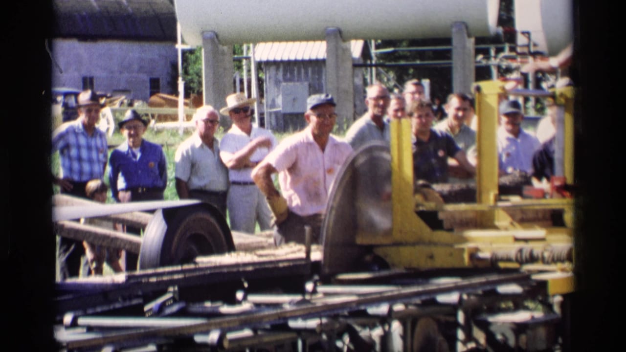 Men watch a yellow wood-cutting machine with a circular saw blade visible in front.