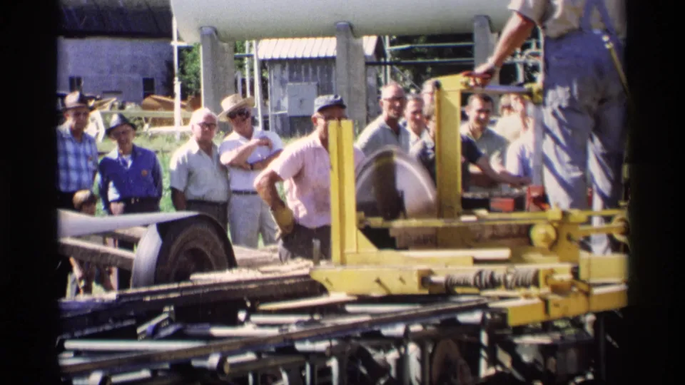 Men in 1969 Mabel, Minnesota observe a yellow wood cutter as an operator’s body is partly visible beside the machinery.