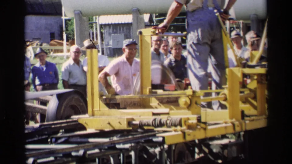 During the 1969 Mabel, Minnesota demonstration, a man in overalls stands on the yellow cutter as observers watch behind him.