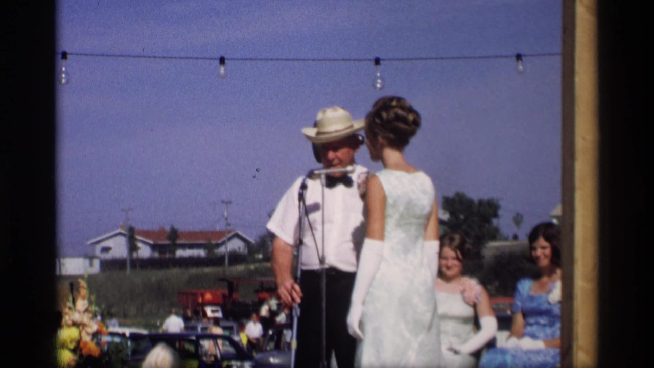 Man in a straw hat speaks at microphones beside a woman in a white dress at an outdoor festival.