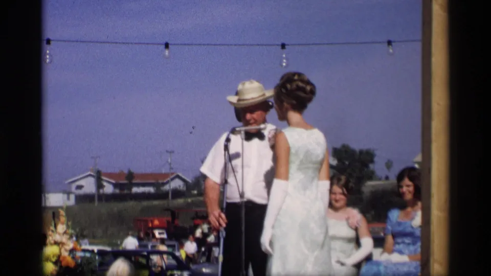 In Mabel, Minnesota, 1969, a man in a straw hat addresses microphones while a woman in a white dress and gloves stands beside him.