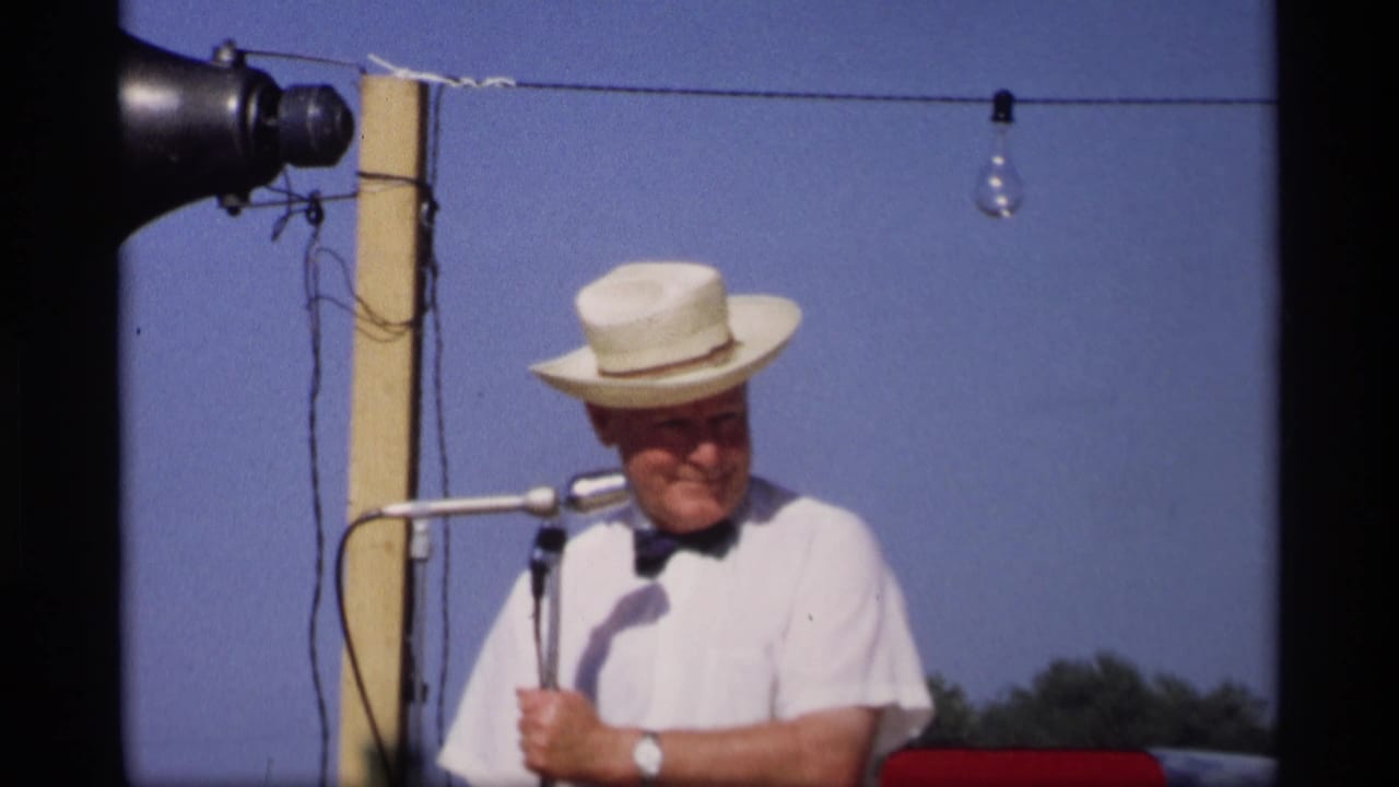 A 1969 Mabel, Minnesota festival host in a straw hat and bow tie stands at paired microphones beneath outdoor string lights.