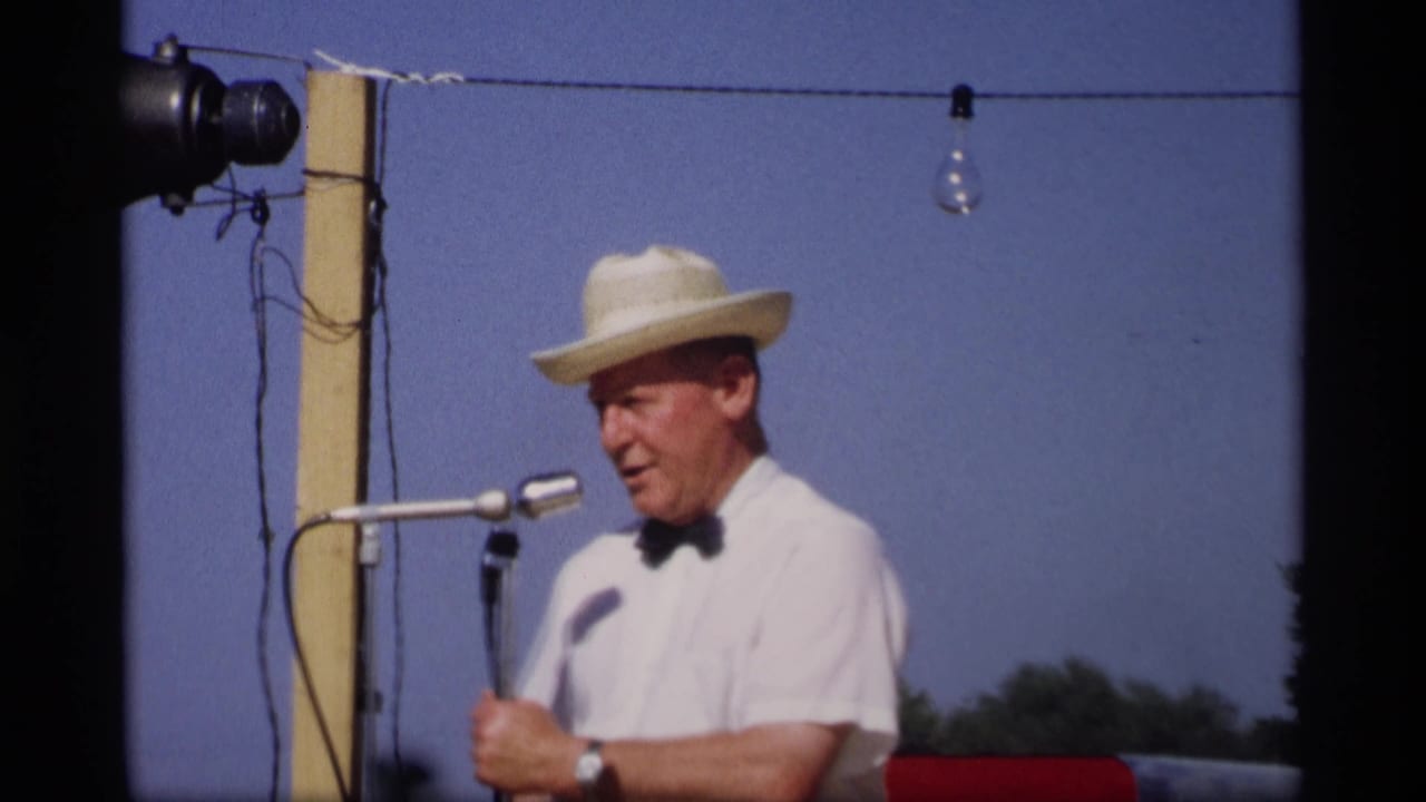 The Mabel, Minnesota festival speaker continues at the microphone, framed close against sky, string lights, and a wooden support pole.