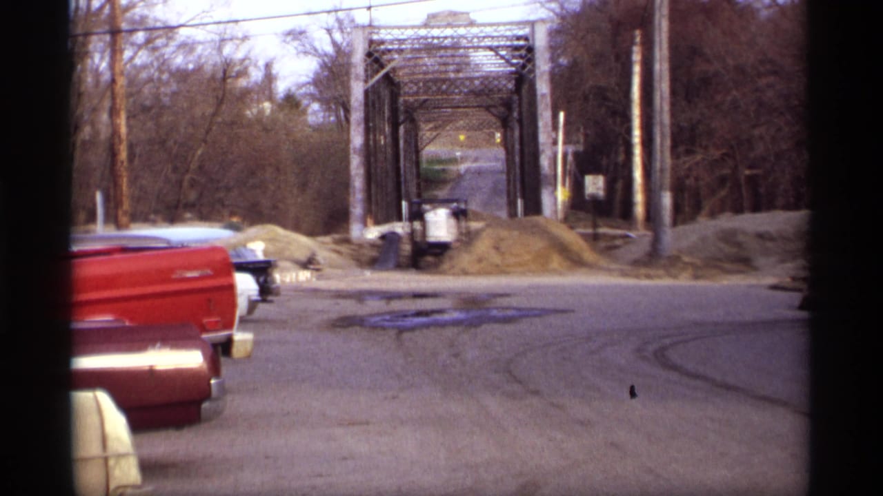 Blue sedan near a metal truss bridge with red pickup trucks along the roadside.