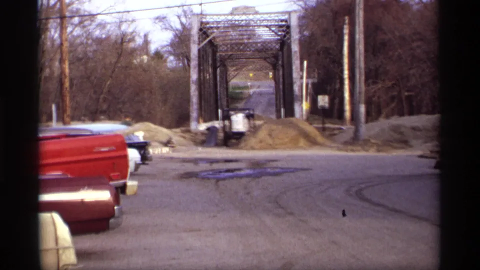 1969 Rockford, Minnesota street scene with a blue classic sedan approaching a metal truss bridge near red pickup trucks and gravel piles.