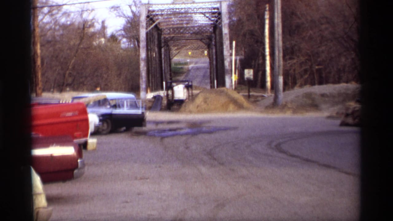 Blue sedan turning near gravel piles and a metal truss bridge.