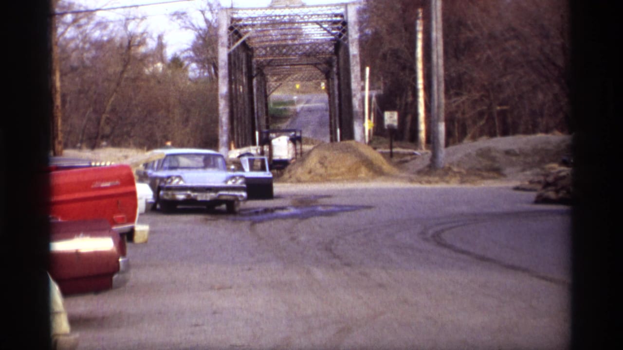 A blue sedan appears stopped with a door open near a red pickup truck and metal truss bridge in Rockford, Minnesota.