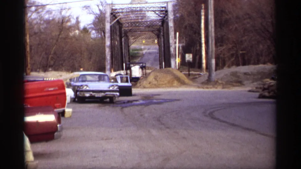 A blue sedan appears stopped with a door open near a red pickup truck and metal truss bridge in Rockford, Minnesota.