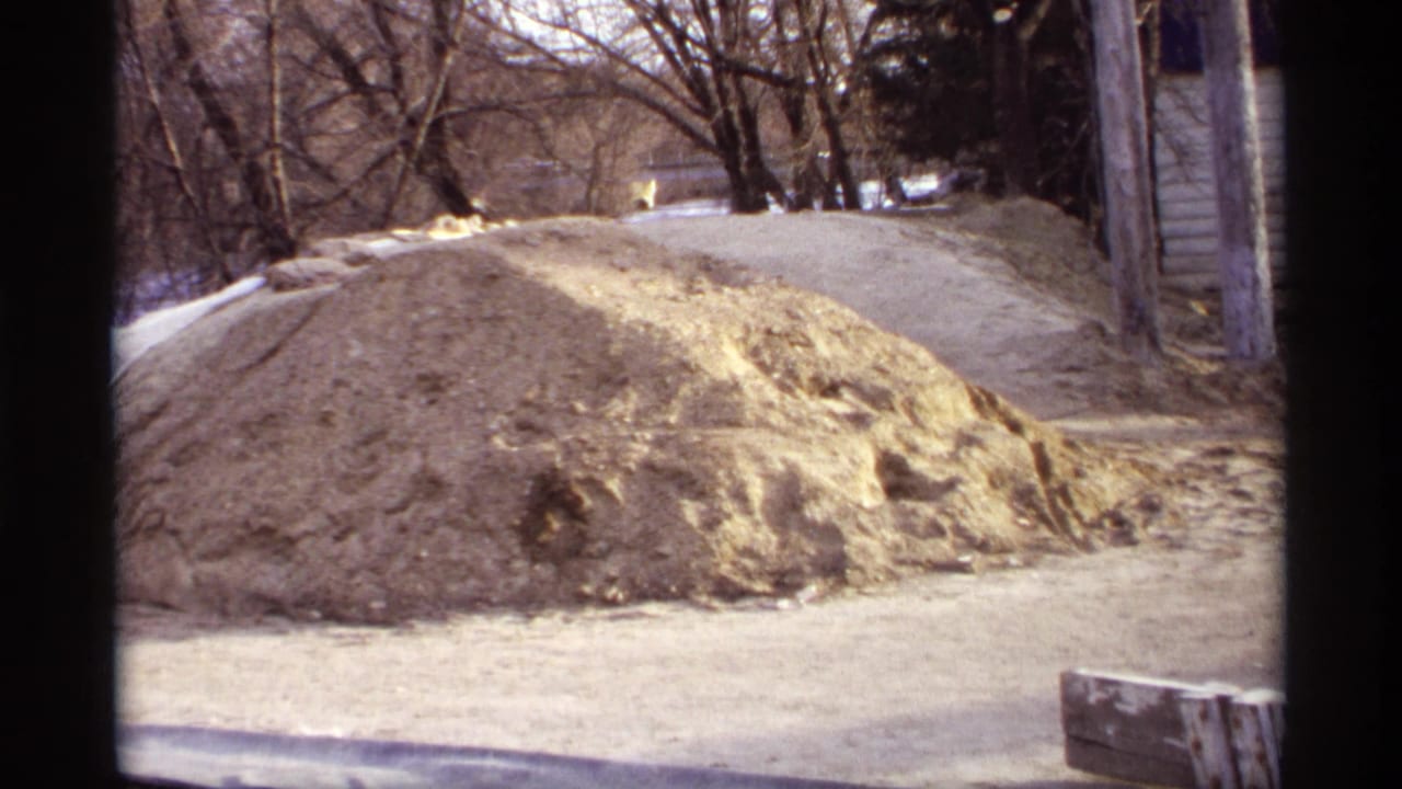 Roadside construction material fills the foreground in a 1969 Rockford, Minnesota home-movie view near trees and a building edge.
