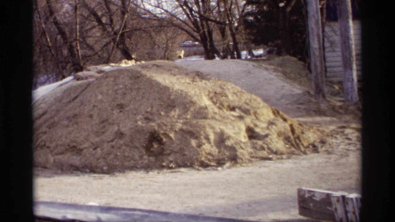 Large sand mound beside a road with bare trees and buildings in the background.