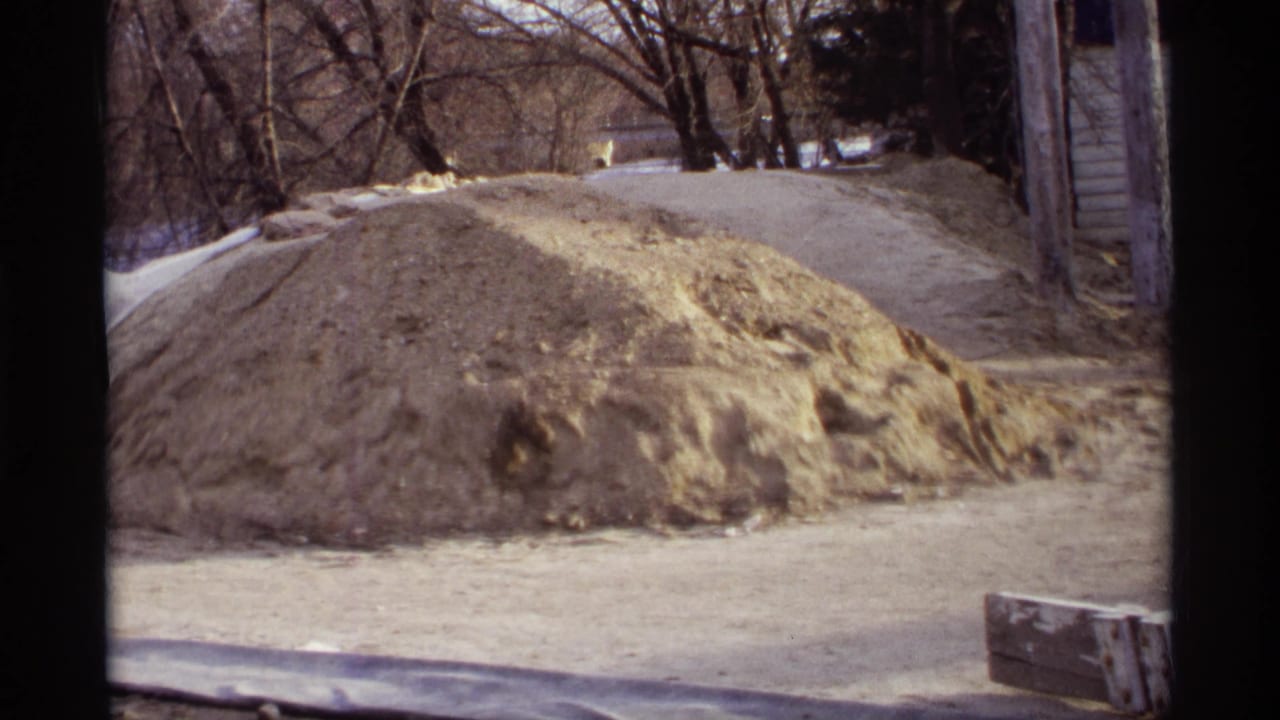 1969 Rockford, Minnesota 8mm view of a sand mound by a road, with bare trees and nearby structures in a flood-defense setting.