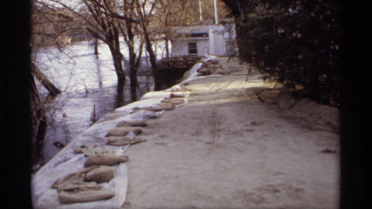 1969 Rockford, Minnesota frame showing sandbags along a raised riverbank beside floodwater, with bare trees and a white building in the background.