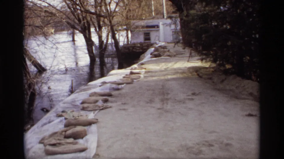 1969 Rockford, Minnesota frame showing sandbags along a raised riverbank beside floodwater, with bare trees and a white building in the background.