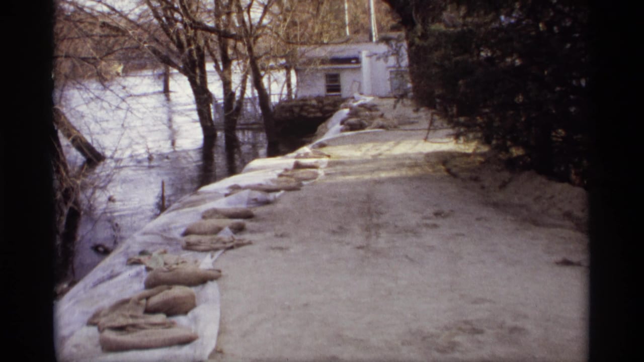 1969 Rockford, Minnesota 8mm frame of a sandbag line beside floodwater, bare trees, and a white building near the riverbank.